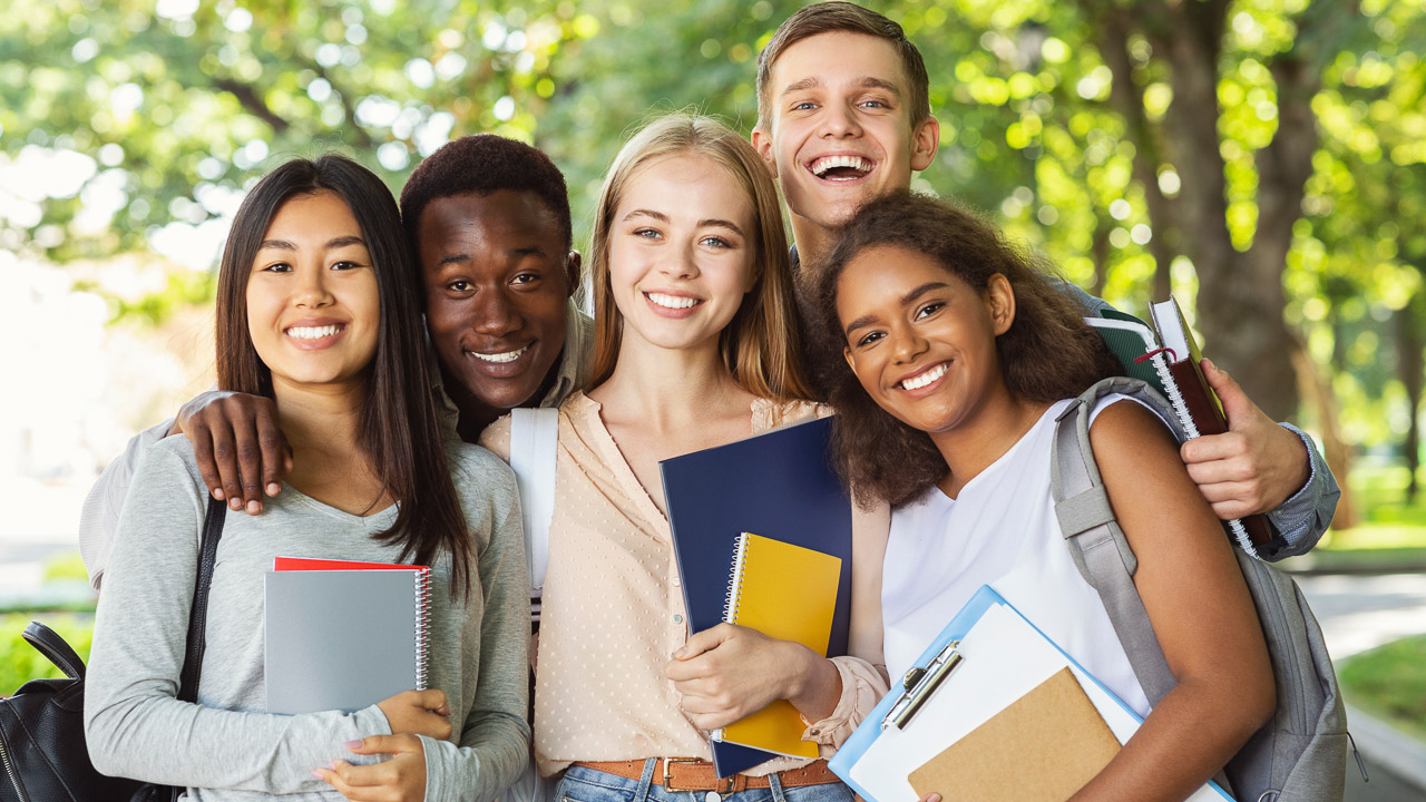 Group of international happy students with books and notebooks having fun in park after studying, smiling at camera